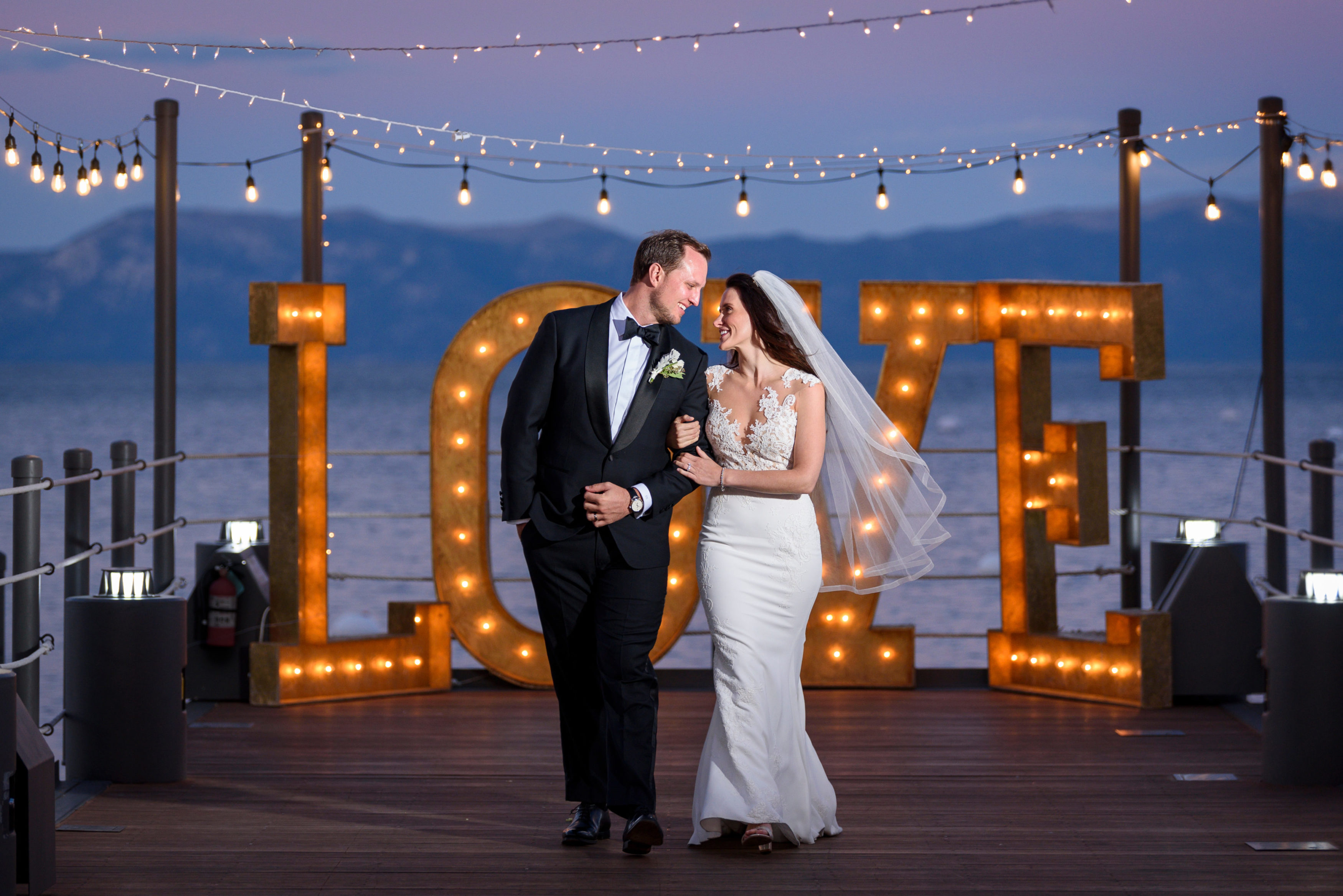 A bride and groom look at each other in front of a large sign that says "Love" on the West Shore Cafe pier over looking Lake Tahoe