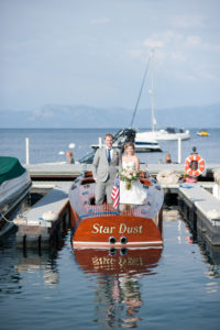 A bride and groom stand on a wooden boat. The words "Star Dust" are painted on the boat. The boat is tied to a dock.