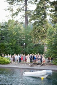 A group of people stand on a lawn outside West Shore Cafe. They appear to be celebrating. A boat floating on a lake is in the foreground. Sandra Fazzino Photography