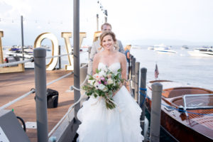 A bride stands on the West Shore Cafe Pier holding a bouquet of flowers. The groom stands behind her. A wooden boat is in the background. Sandra Fazzino Photography