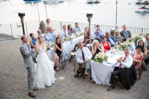 A bride and groom cheers champagne flutes while people sitting at tables in the backgroud applaud. The scene takes place on West Shore Cafe deck overlooking Lake Tahoe