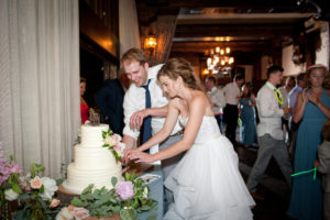 A bride and groom cut a white wedding cake together. People are dancing in the background.