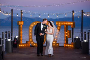 A bride and groom stand in front of a sculpture of the word "Love" on the West Shore Cafe Pier. Lake Tahoe is in the background. Photography by Monique