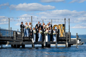 A wedding party stands on the end of West Shore Cafe Pier. The bridesmaids are wearing blue dresses and holding bouquets. The Groomsmen are in suits. Lake Tahoe is in the background.Photography by Monique