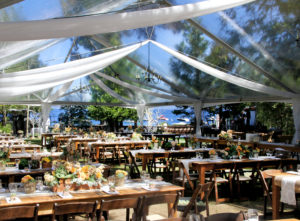 The West Shore Cafe Lawn is set for a dinner reception. There are wooden farm tables with white linens and orange flowers. A large white tent covers the area. Lake Tahoe is in the background.