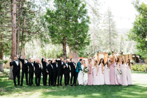 A bride and groom kiss with eight groomsmen in tuxedos on their left and eight bridesmaids in white and pink dresses on the right. They are on the West Shore Cafe LawnCustock Photography