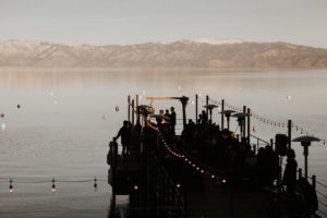 The West Shore Cafe Pier is silhouetted against Lake Tahoe. There is a wedding ceremony on the pier. Snow covered mountains are in the background. Kay Kroshus Photography