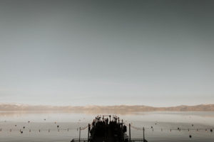 The West Shore Cafe Pier is sillohuetted against Lake Tahoe. A wedding ceremony is in progress.
