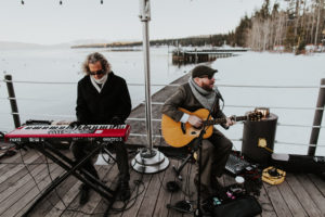 Two men sit on the West Shore Cafe Pier. One is playing a keyboard and the other plays a guitar. There is snow and Lake Tahoe is in the background.